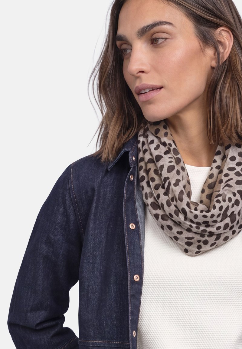 Femme portant une écharpe tachetée marron clair et noire, un haut blanc texturé et une veste en jean bleu foncé, regardant sur le côté sur un fond uni.