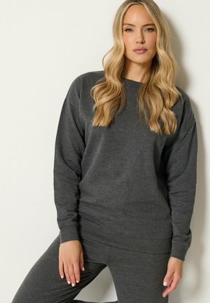Kvinde med langt blondt hår, iført en mørkegrå sweatshirt og matchende bukser, står mod en ensfarvet lys baggrund.