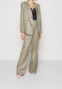Conjunto de blazer metálico dorado y pantalones de pierna ancha, con una textura brillante. Top negro debajo y tacones de punta rosa.