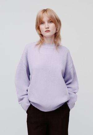 Lila neulepaita, jossa on pyöreä kaula-aukko, alhaiset olkapäät ja ribbauskainalot, yhdistettynä tummiin housuihin. Liukas, pehmeä tekstuuri kauttaaltaan.