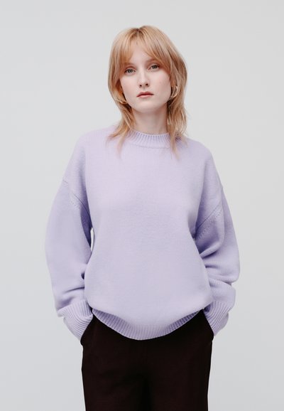 Maglione lavorato a maglia lilla con collo rotondo, spalle scese e polsini a costine, abbinato a pantaloni scuri. Texture liscia e morbida.