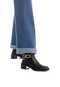 Botas de cuero negras hasta el tobillo con un tacón bajo tipo bloque y detalles en hardware dorado. Aparecen con unos pantalones de denim azul arremangados.