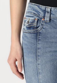 Világoskék farmernadrág réz szegecsekkel, a zsebek körüli látható varrással és sima felülettel. Közeli kép egy kézről, amely a derékon pihen.