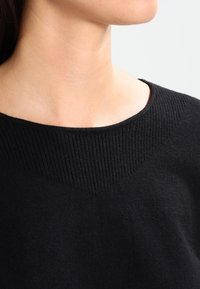 Černý pletený svetr s širokým výstřihem a ribbovanou texturou na ramenou, který zdůrazňuje měkkou tkaninu a minimalistický design.