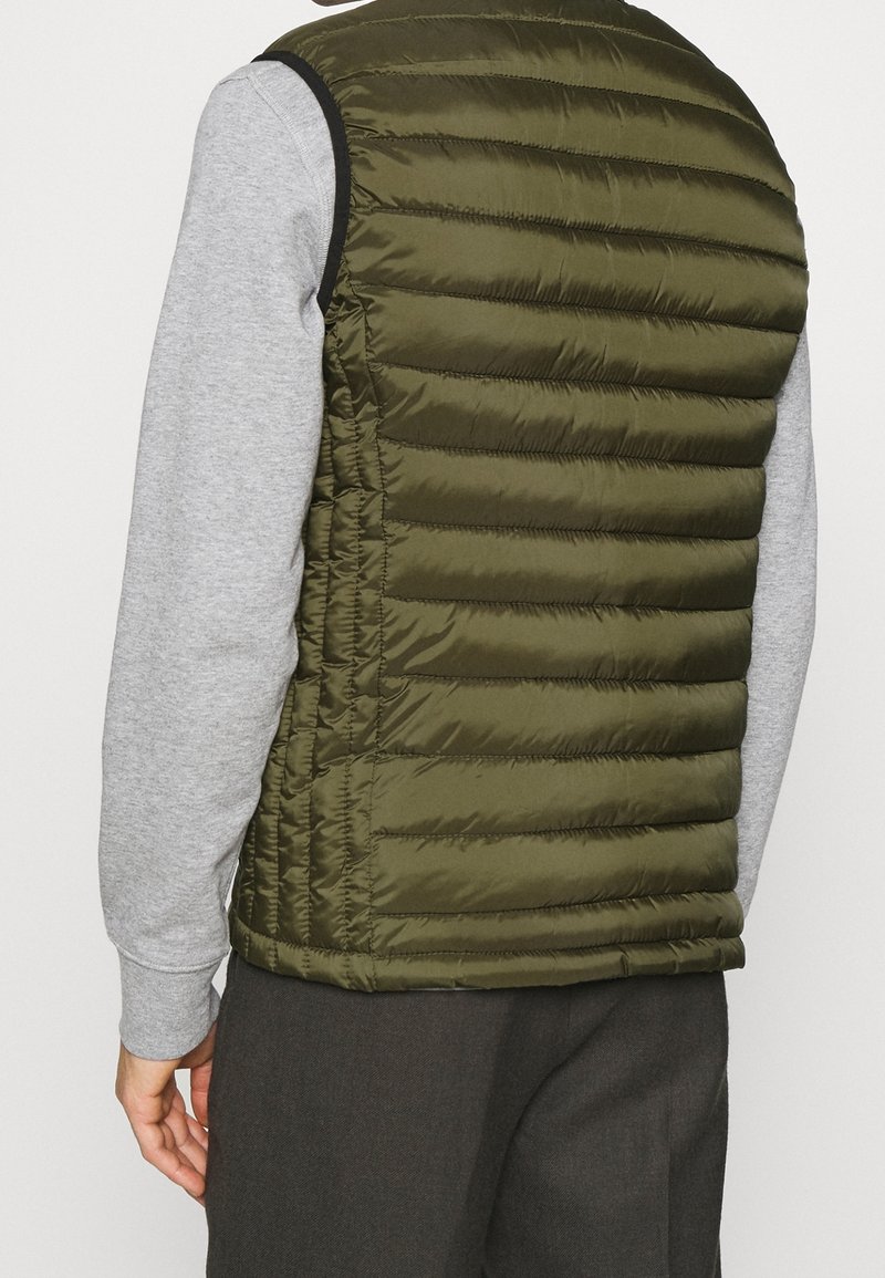 Personne portant un gilet matelassé sans manches vert olive sur une chemise gris clair à manches longues, vue de dos.
