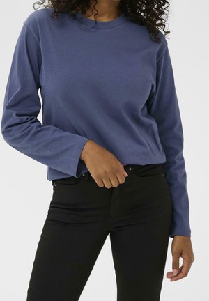 Mujer con una camisa azul de manga larga metida en unos pantalones negros de talle alto, posando con una mano cerca de la cintura.