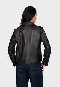 Blouson en cuir noir de moto avec épaulettes, poignets zippés et texture lisse. Associé à un pantalon en denim bleu.
