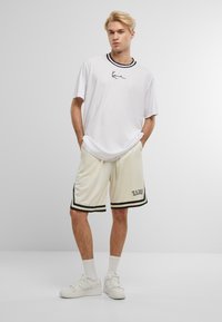 Hvid t-shirt med sort krave og broderet design, parret med cremede shorts med sorte detaljer og "KANI" tekst. Hvide sneakers.