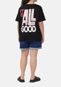 Schwarzes Baumwoll-T-Shirt mit auffälligem weißen und pinken Grafiktext auf dem Rücken, kombiniert mit Jeansshorts und schwarzen Slipper-Schuhen.