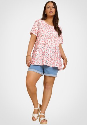 Donna con una maglietta a maniche corte rosa a fiori, pantaloncini di jeans blu con risvolto e sandali bianchi con cinturino alla caviglia, in piedi contro uno sfondo semplice.