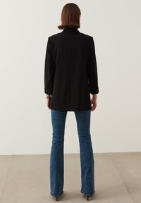 Blazer negro entallado con solapa, mangas enrolladas, combinado con jeans acampanados de color azul. Tejido suave, forma estructurada y detalles mínimos.