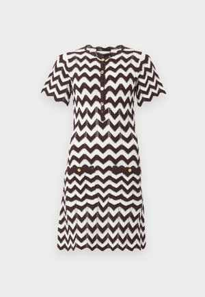 Žena odjevena u kratku haljinu s crno-bijelim cik-cak uzorkom, prednjim džepovima i bijelim slingback cipelama s mašnama.