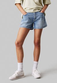 Shorts de mezclilla en azul claro con un ajuste relajado, dobladillo deshilachado, dos bolsillos laterales, combinados con zapatillas blancas y calcetines texturizados.