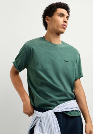 Giovane uomo che indossa una t-shirt verde e una camicia a righe legata intorno alla vita, che guarda di lato contro uno sfondo chiaro e uniforme.