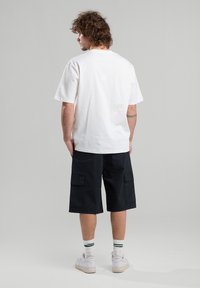 T-shirt en coton blanc à manches courtes, assorti à un short cargo noir avec des poches latérales. Le modèle porte des baskets blanches avec des chaussettes à rayures vertes.