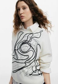 Suéter blanco con un diseño de línea abstracta negra en la parte frontal. Textura de canalé, escote redondo y cuello de camisa blanca superpuesto.
