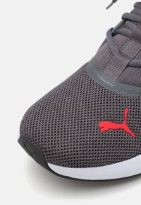 Grå atletiksko med mesh-overdel, rød Puma-logo, polstret tunge og sort gummisål. Har snørebånd og tekstureret overflade.