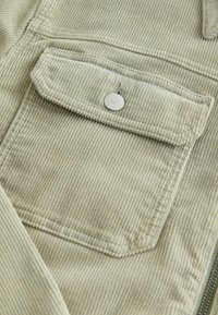 Tissu en velours côtelé vert clair avec une poche à rabat carrée sécurisée par un seul bouton-pression rond en argent.