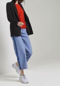 Blazer negro sobre un suéter rojo de canalé, combinado con jeans de pierna ancha en azul claro que tienen una franja lateral azul oscura y zapatillas blancas.