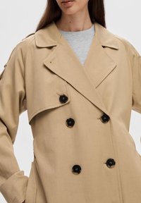 Beige dubbelknäppt trench coat med svarta knappar som bärs över en ljusgrå crew neck-tröja, med armarna i sidan.