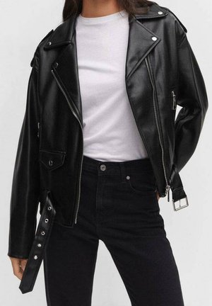 Vrouw draagt een zwarte leren motorjack met rits, gespsluiting en drukknoopdetails over een wit T-shirt en zwarte broek.