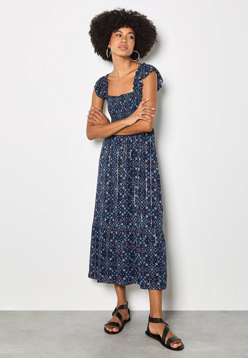 Femme aux cheveux bouclés portant une robe midi bleu marine à motif floral avec des manches à volants et des sandales noires à brides, debout, les bras croisés.