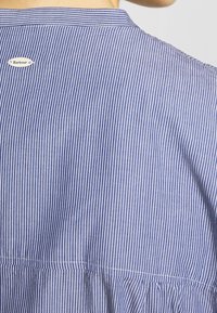 Modrá a bílá pruhovaná košile s límečkem, na zadní straně s kovovým štítkem značky Barbour a s nařasenou spodní částí.