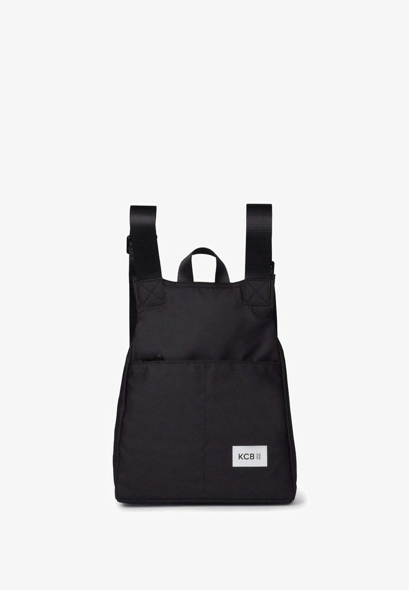 Mochila negra con bolsillo frontal con cremallera, dos correas ajustables para los hombros, asa superior y parche con el logo blanco de KCB en la parte inferior derecha del frente.