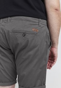 Pantalones cortos grises de tela, con un bolsillo trasero con cierre de botón y una etiqueta de cuero con el logo. Detalle de dobladillo enrollado visible.