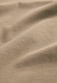 Beige kokvilnas audums ar gludu tekstūru, iekļaujot maigas krokas un smalku spīdumu, demonstrējot mīkstu un vieglu izskatu.