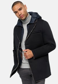 Black coat with a navy hood, featuring a double-breasted design, contrasting trim, and a grey turtleneck. Textured fabric with a tailored fit.