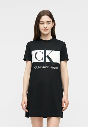 Fiatal nő hosszú barna hajjal, fekete Calvin Klein Jeans pólóruhát visel, melynek elején nagy fehér és fekete logó található.