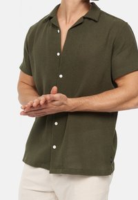 Chemise manches courtes vert olive avec un tissu texturé, col ouvert et boutons blancs ; présente un ourlet droit et une petite étiquette de logo.