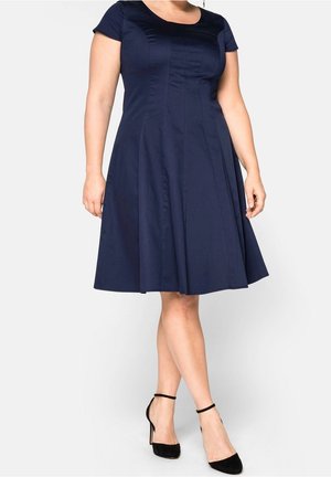 Mujer con vestido ajustado y acampanado de manga corta azul marino y tacones negros con tiras en el tobillo, posando frente a un fondo liso y claro.