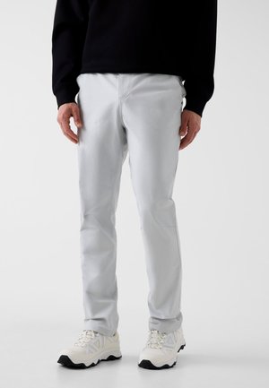Hombre con pantalones gris claro, zapatillas blancas con suelas negras y una camiseta negra de manga larga, de pie contra un fondo liso.
