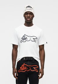 Λευκό βαμβακερό T-shirt με μαύρο σχέδιο ενός πηδώντας γάτου, συνδυασμένο με μαύρο τζιν που έχει σχέδιο πορτοκαλί γάτας στην πίσω τσέπη.