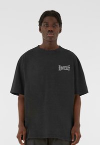 Sort oversized bomuld t-shirt med korte ærmer, der har hvid tekst på venstre side. Stoffet har en blød, let struktureret overflade.