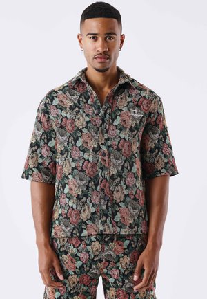 Man draagt een bijpassend kortarmig overhemd en broek met een meerkleurig bloemenpatroon op een lichte achtergrond.