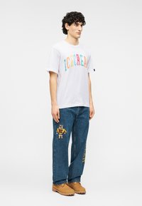 Λευκό T-shirt με πολύχρωμο κείμενο "ICECREAM", συνδυασμένο με μπλε τζιν τζιν που έχει γραφικό αρκούδας και κίτρινες λεπτομέρειες. Καφέ παπούτσια.