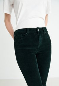 Pantalon en velours côtelé vert foncé avec une texture côtelée, doté de poches avant et d'une fermeture à bouton, associé à une chemise blanche.