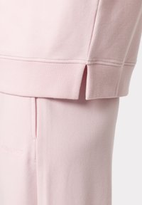 Světle růžový top z bavlněné směsi pletený s žebrovaným lemem a bočními rozparky, s jemným logem v reliéfním textu poblíž spodní části.