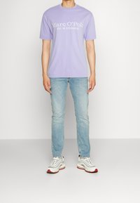 T-shirt à manches courtes lavande avec le texte blanc "Marc O’Polo EST. À STOCKHOLM," associé à un jean bleu clair et des baskets multicolores.