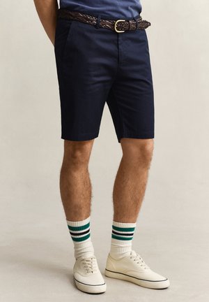 Mann iført marineblå shorts, vevd brun belte, hvite joggesko og hvite crew-sokker med grønne og svarte striper, stående på ensfarget bakgrunn.