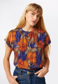 Kortærmet bluse med livligt orange og blåt blomsterprint, knappedetaljer og indsamlet halsudskæring. Båret med denimjeans.