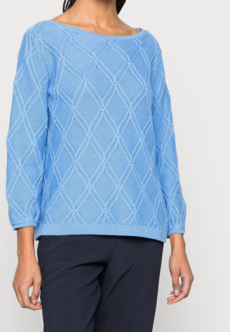 Nainen, joka on pukeutunut vaaleansinisessä puuvillaneuleessa, jossa on timanttikuviota, ja mustissa housuissa, seisoo tasaisella taustalla.