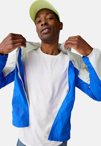 Helle blaue und weiße, leichte Jacke mit Reißverschluss vorne, glatter Textur und elastischen Bündchen. Über einem weißen Hemd getragen.