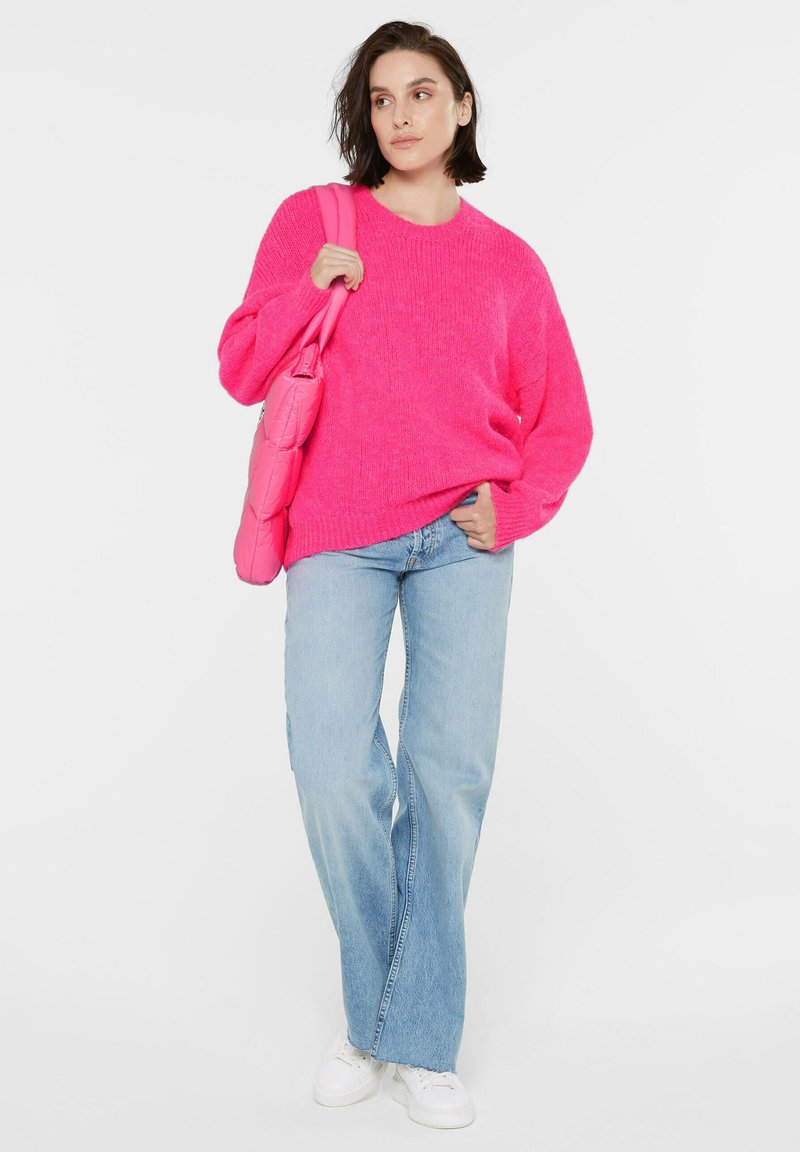 Helles, pinkes, übergroßes Strickpullover mit langen Ärmeln, kombiniert mit hellblauen Schlagjeans und weißen Sneakers. Akzentuiert mit einer pinken Tasche.