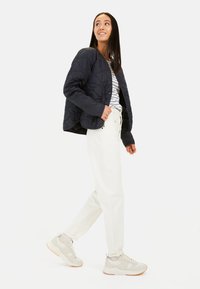 Marineblau gesteppte Jacke mit Rundhalsausschnitt, kombiniert mit einem gestreiften Hemd und weißen, gerade geschnittenen Hosen; hellgraue Sneakers mit orangefarbigen Sohlenakzenten.