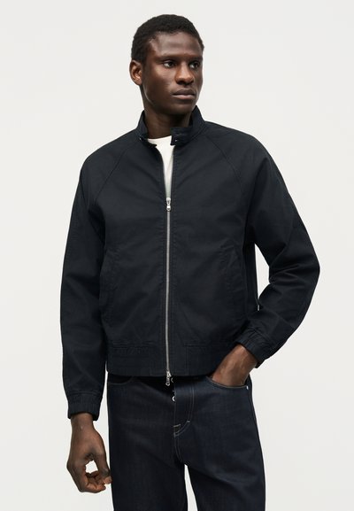 Άνδρας που φοράει μαύρο μπουφάν με φερμουάρ, λευκό πουκάμισο από κάτω και σκούρο τζιν, στέκεται με το ένα χέρι στην τσέπη σε μονόχρωμο φόντο.