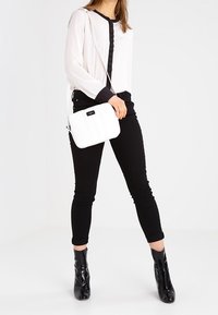 Kvinna i svarta anklarboots, svarta byxor, vit blus med svarta detaljer, som bär en vit axelväska mot en vit bakgrund.